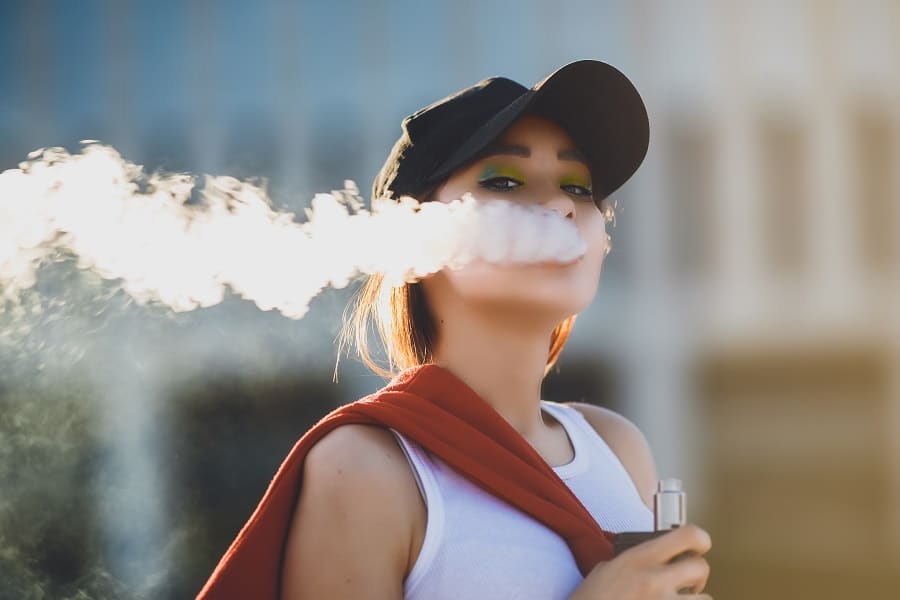 Pretty young hipster asian woman in black hat vape ecig, vaping device at the sunset. Toned image. Closeup