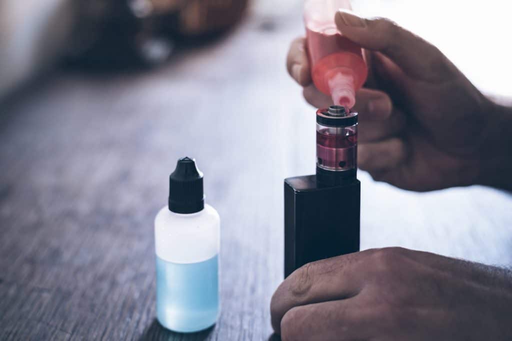 hands of a male person refilling the tank of an e-cigarette with red liquid
