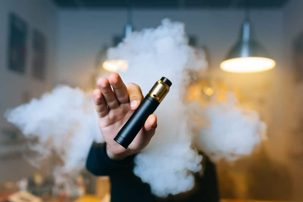 Young beard man show vaping device on his outstretched hand through a cloud of steam. Selective focus. Vaping concept. Copy space