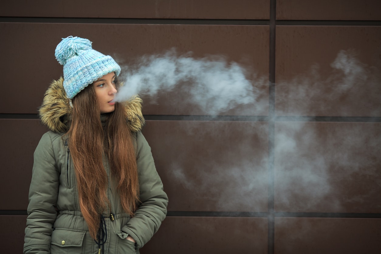 Vape teenager. Young pretty white girl smoking an electronic cigarette opposite modern brown background on the street in the winter.