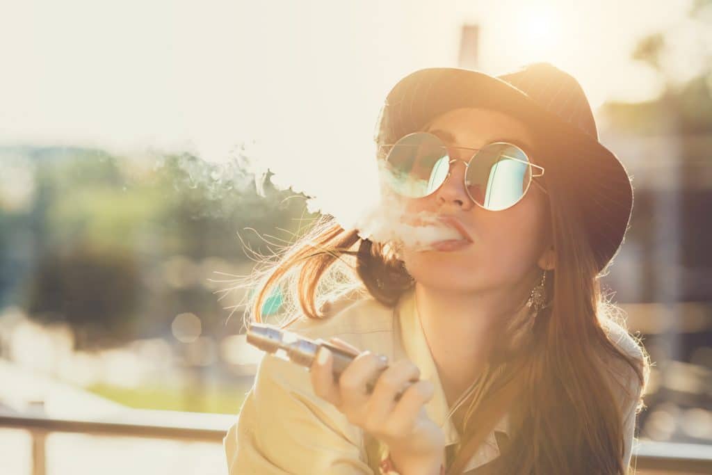 Pretty young hipster woman in black hat vape ecig, vaping device at the sunset. Toned image