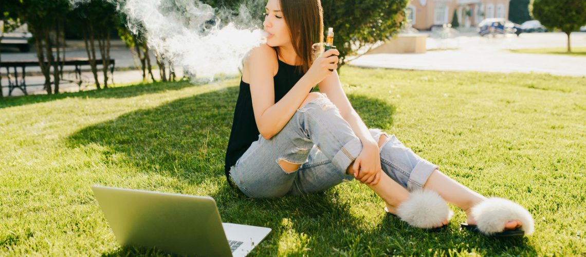 Outdoors photo of trendy attractive girl smoking vape, spending leisure time while sitting on grass in the park, with laptop.