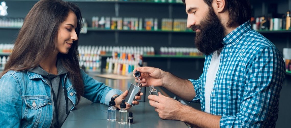 The seller in the vipeshop shows how to fill an electronic cigarette. He is a tall man with long hair and a beard. The store has a large selection of of electronic cigarettes.