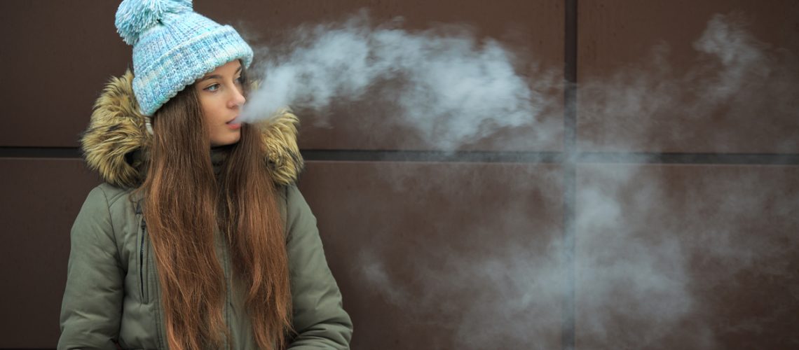 Vape teenager. Young pretty white girl smoking an electronic cigarette opposite modern brown background on the street in the winter.